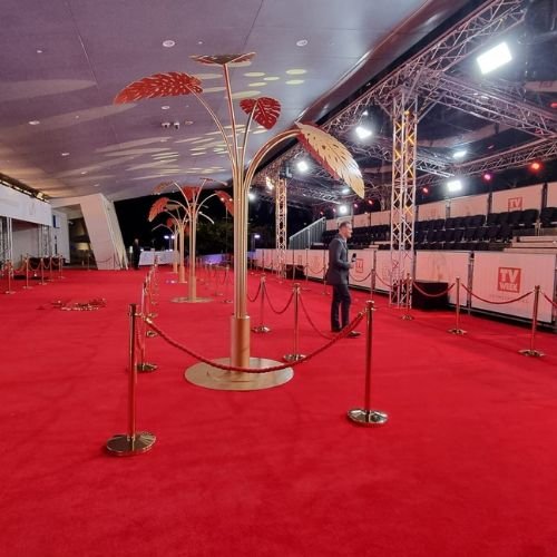 a-wide-red-carpet-event-area-with-gold-stanchions-red-rope-barriers-and-tall-decorative-gold-palm-leaf-sculptures-under-an-outdoor-covered-venue